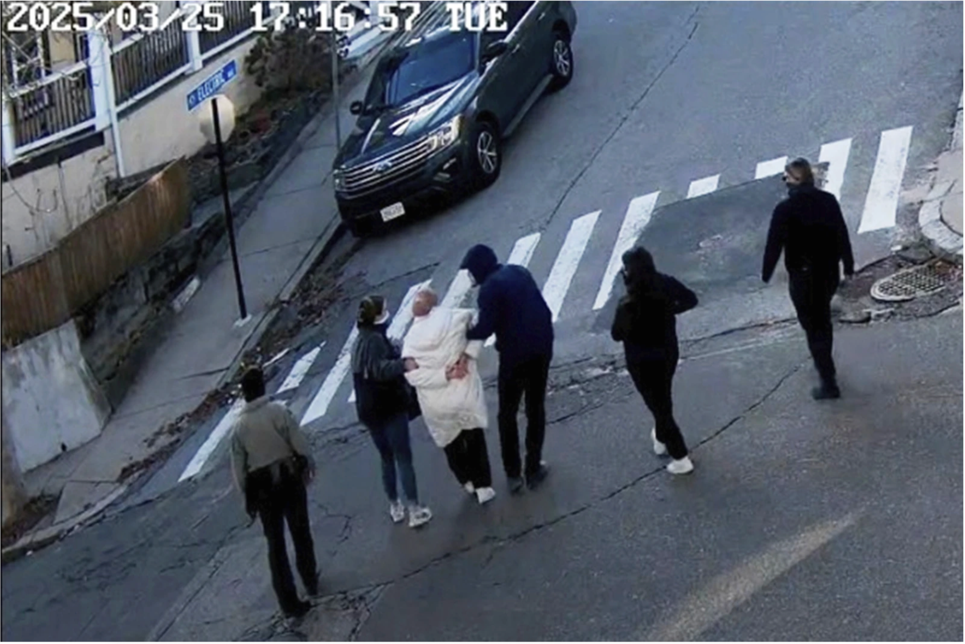 Rumeysa Ozturk, in a white coat and handcuffs, is led towards an ummarked black vehicle by five figures.